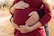 Two pairs of hands gently rest on a pregnant belly, symbolizing care and expectation. The person is wearing a red sweater, and there are wedding rings visible on some of the fingers. The background is blurred, with hints of autumnal colors.