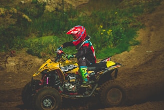A person wearing colorful protective gear and a helmet rides an ATV on a dirt track surrounded by greenery. The ATV is predominantly yellow with various decals.
