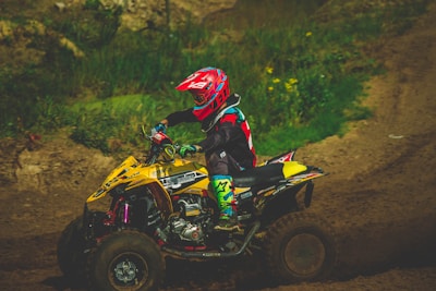 A person wearing colorful protective gear and a helmet rides an ATV on a dirt track surrounded by greenery. The ATV is predominantly yellow with various decals.