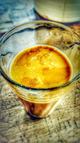 A glass filled with a creamy, frothy espresso shot sitting on a wooden table. The crema is rich and golden, with a slightly marbled pattern across the surface.