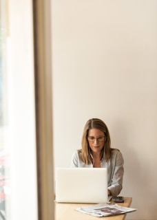 woman wearing grey striped dress shirt sitting down near brown wooden table in front of white laptop computer