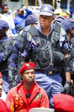 A group of uniformed police officers are standing, with a prominent officer wearing protective gear in the center. In front of him sits a man in a red beret and red clothing, looking serious. The scene appears outdoors with several people visible in the blurry background.