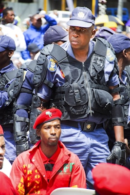 A group of uniformed police officers are standing, with a prominent officer wearing protective gear in the center. In front of him sits a man in a red beret and red clothing, looking serious. The scene appears outdoors with several people visible in the blurry background.