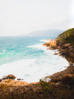 A serene coastal landscape with turquoise waves crashing against rugged cliffs. The rocks have a golden-brown hue and are partially covered with green vegetation. The horizon is marked by a faint outline of distant hills beneath a light, overcast sky.