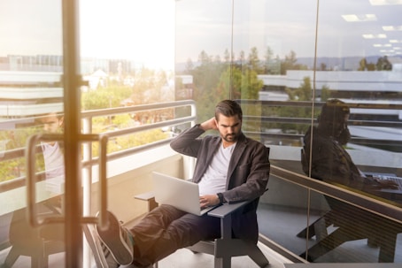 man sitting on gray arm chair using silver laptop computer on building balcony at daytime
