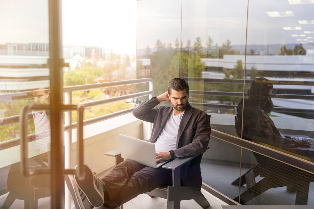 man sitting on gray arm chair using silver laptop computer on building balcony at daytime