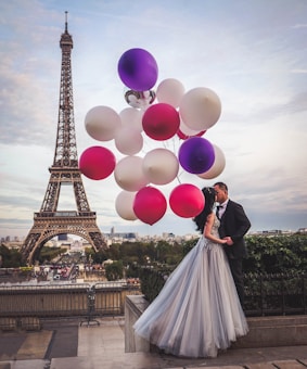 A couple is embracing near the Eiffel Tower, with the man in a suit and the woman in a flowing gown. They are surrounded by a cluster of colorful balloons, primarily pink, white, and purple. The Eiffel Tower and a view of Paris are visible in the background.