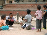 A group of children gather outdoors on a brick surface near a historic structure with chains and stone carvings in the background. They are engaged in conversation, with some sitting and others standing, appearing focused and jovial. Bright clothing and varied postures contribute to a lively atmosphere.