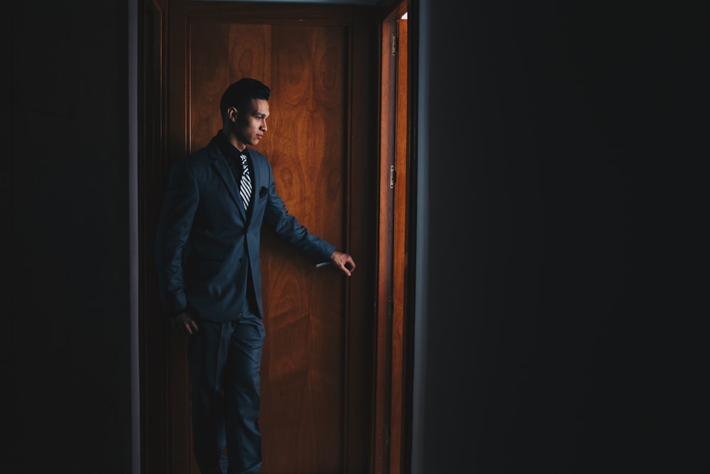 man wearing black suit jacket and dress pants standing beside brown wooden door