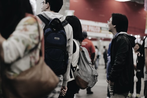 A group of people stand in a queue, each carrying backpacks and shoulder bags. The setting appears to be indoors, possibly an event or a busy public space with a red wall in the background. The people are dressed casually and focus their attention in one direction, likely waiting or engaged in observing something.