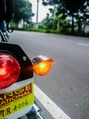 The image displays a close-up of a motorcycle's rear section featuring a red taillight and an orange indicator light. A license plate with text is partially visible. The background includes a road and blurred greenery, suggesting a roadside environment.