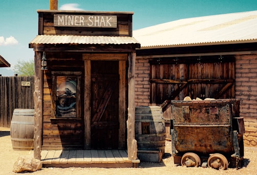 A rustic, wooden shack labeled 'Miner Shak' sits in a dusty, old-west setting. The structure is flanked by large wooden barrels, and an old metal mining cart filled with rocks is positioned to the right. The surroundings include a wooden fence and a brick building, under the bright and clear sky.