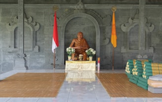 A statue of a monk is seated on a pedestal in a decorative alcove with ornate carvings. To the left and right of the statue are two flags—one red and white, the other orange with a circular emblem. Floral arrangements are placed on either side of the statue, and several folded green and gold mats or cushions are stacked to the right.