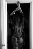 A person stands with their back to the camera, arms raised, wearing sheer, lacy black lingerie in a doorway. The environment is dimly lit, adding shadows and highlights to the contours of the person's body.