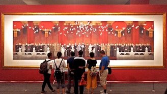 A large painting is displayed on a red wall, featuring numerous people dressed in formal attire, positioned in a grand architectural setting with prominent red columns and hanging lanterns. White doves are depicted flying above the group. In front of the painting, several viewers stand closely examining it.