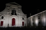 A historic church building illuminated at night, with distinctive architectural features including ornate carvings and red doors. Adjacent to the church is a more modern building, similarly lit, with rectangular windows.