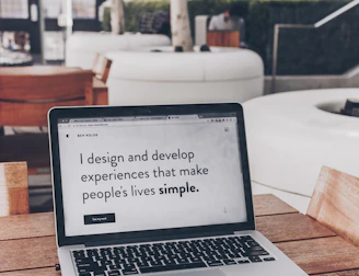 A laptop displaying a webpage with the text 'I design and develop experiences that make people's lives simple.' is placed on a wooden table. It is set in an outdoor seating area with white furniture and greenery in the background. The setting appears to be calm and conducive to work or relaxation.