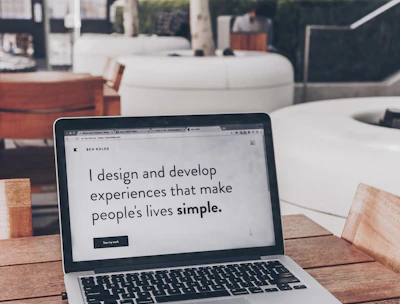 A laptop displaying a webpage with the text 'I design and develop experiences that make people's lives simple.' is placed on a wooden table. It is set in an outdoor seating area with white furniture and greenery in the background. The setting appears to be calm and conducive to work or relaxation.