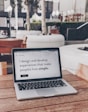 A laptop displaying a webpage with the text 'I design and develop experiences that make people's lives simple.' is placed on a wooden table. It is set in an outdoor seating area with white furniture and greenery in the background. The setting appears to be calm and conducive to work or relaxation.