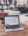 A laptop displaying a webpage with the text 'I design and develop experiences that make people's lives simple.' is placed on a wooden table. It is set in an outdoor seating area with white furniture and greenery in the background. The setting appears to be calm and conducive to work or relaxation.