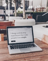 A laptop displaying a webpage with the text 'I design and develop experiences that make people's lives simple.' is placed on a wooden table. It is set in an outdoor seating area with white furniture and greenery in the background. The setting appears to be calm and conducive to work or relaxation.