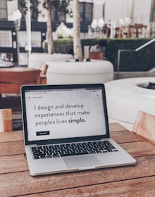 A laptop displaying a webpage with the text 'I design and develop experiences that make people's lives simple.' is placed on a wooden table. It is set in an outdoor seating area with white furniture and greenery in the background. The setting appears to be calm and conducive to work or relaxation.