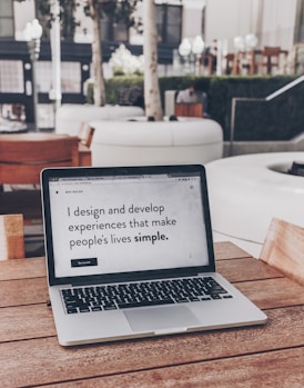 A laptop displaying a webpage with the text 'I design and develop experiences that make people's lives simple.' is placed on a wooden table. It is set in an outdoor seating area with white furniture and greenery in the background. The setting appears to be calm and conducive to work or relaxation.