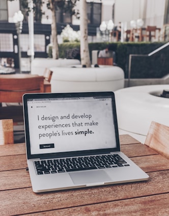 A laptop displaying a webpage with the text 'I design and develop experiences that make people's lives simple.' is placed on a wooden table. It is set in an outdoor seating area with white furniture and greenery in the background. The setting appears to be calm and conducive to work or relaxation.