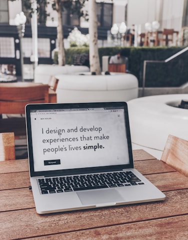 A laptop displaying a webpage with the text 'I design and develop experiences that make people's lives simple.' is placed on a wooden table. It is set in an outdoor seating area with white furniture and greenery in the background. The setting appears to be calm and conducive to work or relaxation.
