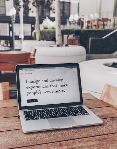A laptop displaying a webpage with the text 'I design and develop experiences that make people's lives simple.' is placed on a wooden table. It is set in an outdoor seating area with white furniture and greenery in the background. The setting appears to be calm and conducive to work or relaxation.