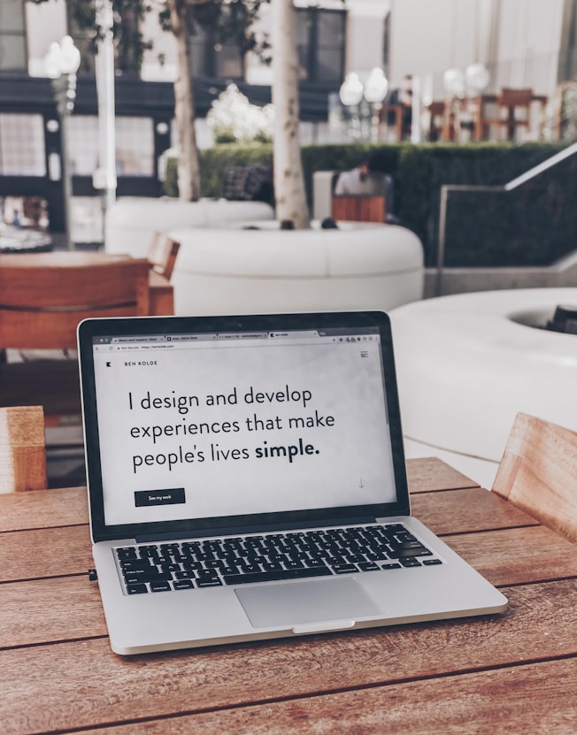 A laptop displaying a webpage with the text 'I design and develop experiences that make people's lives simple.' is placed on a wooden table. It is set in an outdoor seating area with white furniture and greenery in the background. The setting appears to be calm and conducive to work or relaxation.