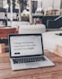 A laptop displaying a webpage with the text 'I design and develop experiences that make people's lives simple.' is placed on a wooden table. It is set in an outdoor seating area with white furniture and greenery in the background. The setting appears to be calm and conducive to work or relaxation.