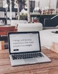 A laptop displaying a webpage with the text 'I design and develop experiences that make people's lives simple.' is placed on a wooden table. It is set in an outdoor seating area with white furniture and greenery in the background. The setting appears to be calm and conducive to work or relaxation.