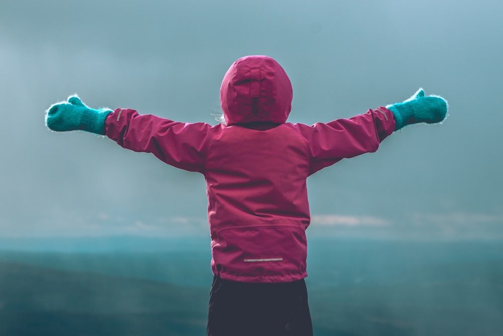 person wearing pink hooded jacket raising her hand in front of green mountain range during daytime