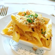 A square white dish contains creamy pasta with a breadcrumb topping, garnished with chopped chives. The dish rests on a lace doily placed on a white plate, with a fork positioned in the background.