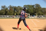 A softball player wearing a blue and red uniform is preparing to hit a pitch on a sunny day. The player is holding the bat in a ready position, and the ball is approaching in mid-air. In the background, another player is visible on the field, and trees border the area beyond a fence.
