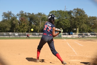 A softball player wearing a blue and red uniform is preparing to hit a pitch on a sunny day. The player is holding the bat in a ready position, and the ball is approaching in mid-air. In the background, another player is visible on the field, and trees border the area beyond a fence.
