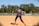 A softball player wearing a blue and red uniform is preparing to hit a pitch on a sunny day. The player is holding the bat in a ready position, and the ball is approaching in mid-air. In the background, another player is visible on the field, and trees border the area beyond a fence.