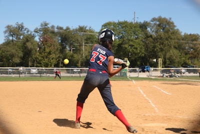 A softball player wearing a blue and red uniform is preparing to hit a pitch on a sunny day. The player is holding the bat in a ready position, and the ball is approaching in mid-air. In the background, another player is visible on the field, and trees border the area beyond a fence.