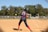 A softball player wearing a blue and red uniform is preparing to hit a pitch on a sunny day. The player is holding the bat in a ready position, and the ball is approaching in mid-air. In the background, another player is visible on the field, and trees border the area beyond a fence.