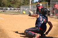 A catcher in full gear is in a crouched position on a baseball or softball field, reaching out to catch a ball mid-air. The scene is set on a sunny day with trees in the background and several people, including players and an umpire, visible near the fence lining the field.