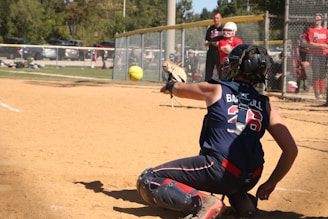A catcher in full gear is in a crouched position on a baseball or softball field, reaching out to catch a ball mid-air. The scene is set on a sunny day with trees in the background and several people, including players and an umpire, visible near the fence lining the field.
