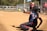 A catcher in full gear is in a crouched position on a baseball or softball field, reaching out to catch a ball mid-air. The scene is set on a sunny day with trees in the background and several people, including players and an umpire, visible near the fence lining the field.