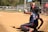 A catcher in full gear is in a crouched position on a baseball or softball field, reaching out to catch a ball mid-air. The scene is set on a sunny day with trees in the background and several people, including players and an umpire, visible near the fence lining the field.