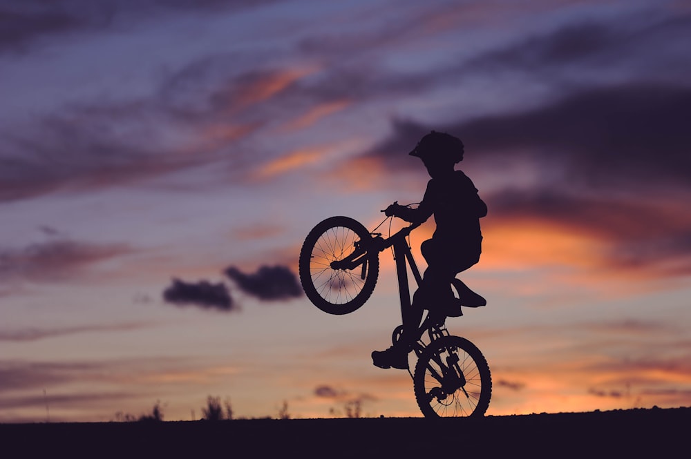silhouette of boy riding bicycle in wheelie during golden hour
