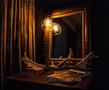A dimly lit room features a wooden-framed mirror reflecting a warm glow from two vintage-style enclosed light bulbs. Below the mirror, on a wooden table, rests a large piece of antler decor alongside a few magazines, one of which has the title 'Frankie' visible. Heavy curtains with a textured, vertical pattern add to the cozy ambiance.