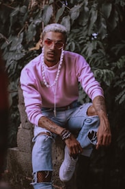 A person with a trendy appearance, wearing a pink sweatshirt and mirrored sunglasses, poses in front of lush green foliage. Their jeans are ripped and they have tattoos on their arms and hands, conveying a stylish, edgy look. Accessories include a white beaded necklace, bracelets, and white sneakers.