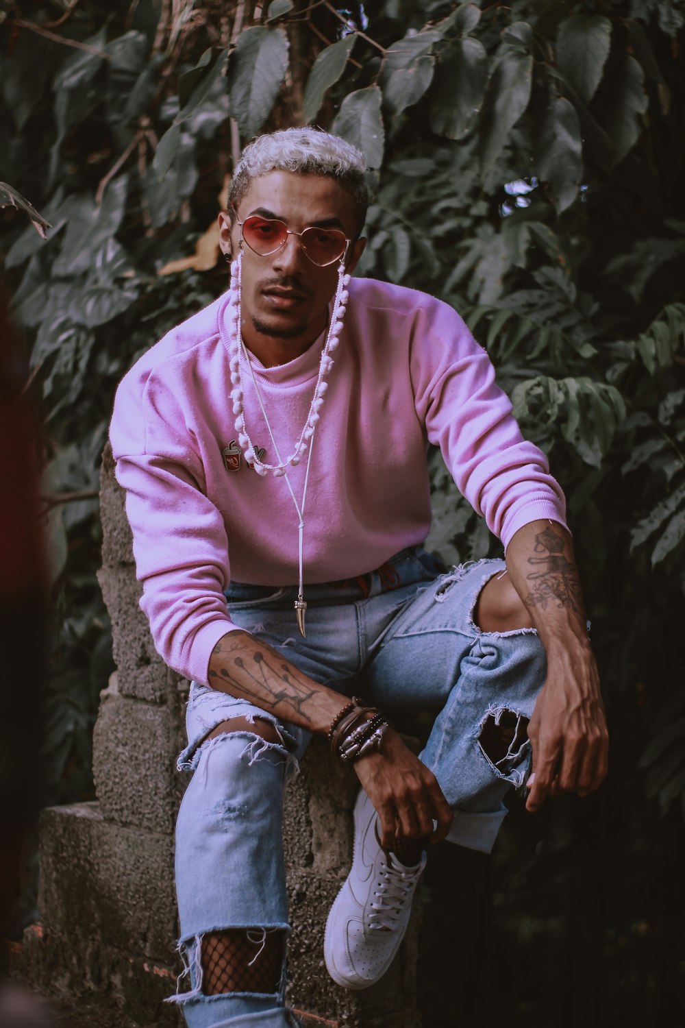 man in pink sweater and distressed blue denim jeans sitting on grey cinder blocks near trees closeup photography