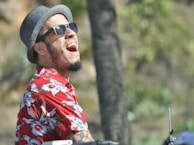 A person wearing sunglasses and a gray hat is joyfully laughing. They are dressed in a vibrant red floral shirt, suggesting a relaxed and tropical setting. The background is blurred with green tones, indicating outdoor surroundings.