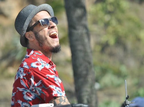 A person wearing sunglasses and a gray hat is joyfully laughing. They are dressed in a vibrant red floral shirt, suggesting a relaxed and tropical setting. The background is blurred with green tones, indicating outdoor surroundings.